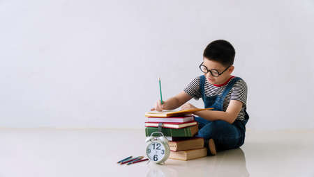 Little cute boy wear eyeglasses sitting on the floor with stack of books , pencil and clock while written on the book with copy space. Schoolboy drawing of homework at home. Black to school concept.の写真素材