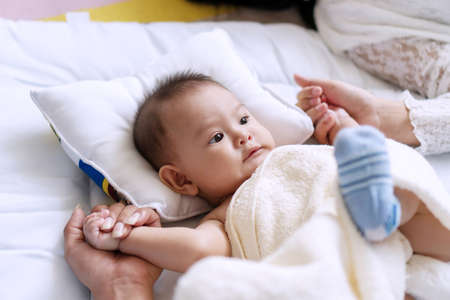 Adorable little baby boy lying on the white bed while holding his parenthood in hands at home. Infant joy kid looking at camera with his father and mother hands. Family relationship concept.の写真素材