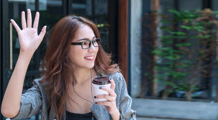 Attractive young woman sitting outdoor in cafÃ© while waving hand saying hello her friend. Business woman wear glasses holding cup of coffee while waving hands greeting her friend say hi or goodbye.の写真素材