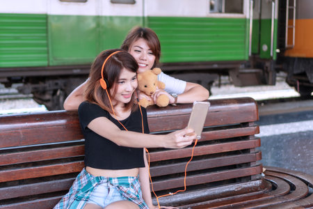 Traveler two young women using smartphone selfie picture to social media in the station. Cheerful girl wear orange earphone with her friend taking photo together while wait train. Friendship concept.の写真素材
