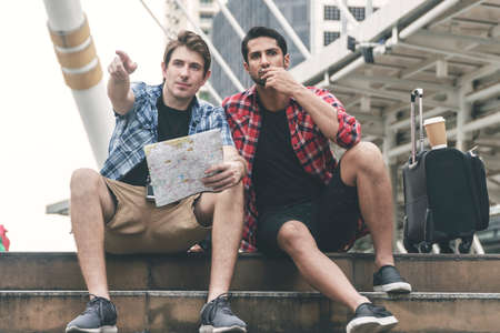 Two traveller handsome guys searching for direction using map paper while sitting on stairs in city. Tourist young man using map on vacation pointing for some destinations. Travel lifestyle concept.の写真素材
