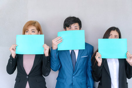 Group of young business woman and businessman wear suit holding green empty sheets of paper and covering half of their faces while looking up something standing on gray background.の写真素材