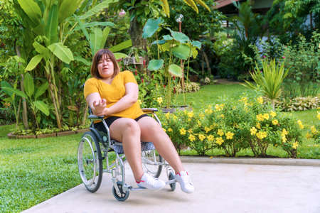 Painful overweight young woman sitting alone on the wheelchair while her hands are sinking due to nervous system illness, hemiplegia and paralysis in the garden. Healthy concept.の写真素材