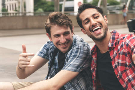 Two handsome friends taking selfie together with action camera sitting on street at city looking the camera making enjoyment face. Two happiness traveller man take picture while travel in holiday.の写真素材