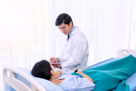 Hospitalized young woman lying in bed while doctor checking her healthcare. Friendly man physician examining female patient in hospital room ward while holding clipboard report in his hand.の写真素材