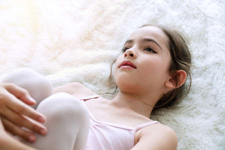 Adorable cute little ballet girl brown long hair lying on white fur rug while looking at camera ....