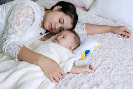 Innocence infant asleep lying on the bed with his mother hug in the bedroom. Young mom cuddling her son sleeping together on the bed after breastfeeding. Parenthood concept.の写真素材