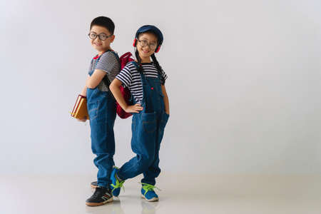 Cheerful brother and sister wear eyeglasses standing back to back together on white background. Little boy hold books in his hand and little girl smile wear earphones with hat. Back to school concept.の写真素材