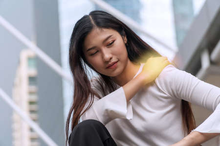 Upset young woman with shoulder pain or stiffness while sitting on city background. Businesswoman overwork tired while her hand touch neck problem stress after work office. Health care concept.の写真素材
