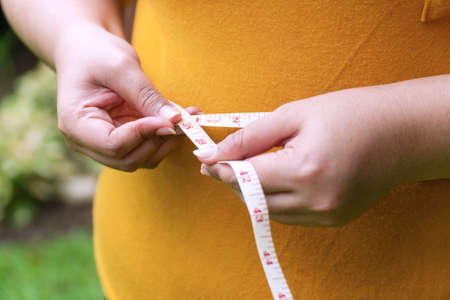 Hand fat woman holding measuring tape around her waist. Overweight female measuring her waist with measuring tape while standing in the garden. Healthcare concept.の写真素材