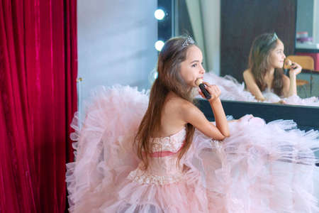 Ballerina little cute girl wear pink dress sitting on a chair in the dressing room and make yourself a makeover. She holding brush in her hand for applying makeup with reflection in mirror.の写真素材