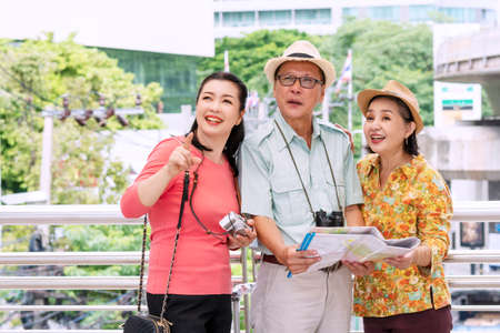 Group of traveler elderly people wear hat with luggage, camera and binoculars holding map in hands while walking together in street city. They are using map on vacation pointing for some destinations.の写真素材