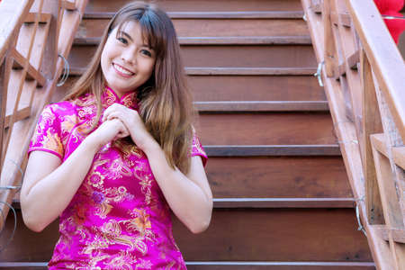 Enjoy young woman wearing pink cheongsam or qipao sitting on stair while raising her hand to bless her to be happy and healthy. she looking at camera with smile. Chinese new year concept.の写真素材