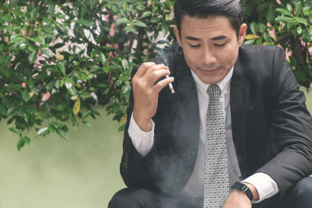 Handsome young businessman wear black suit smoking and thinking about business issues while sitting at outdoor. Young man holding cigarette in his hands while looking at something.の写真素材
