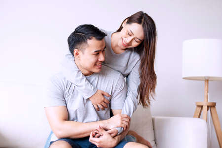 Young woman hugging her boyfriend from back while sitting together on sofa at home. Romantic couple embracing feeling relax in living room. Relationship and valentines day concept.の写真素材