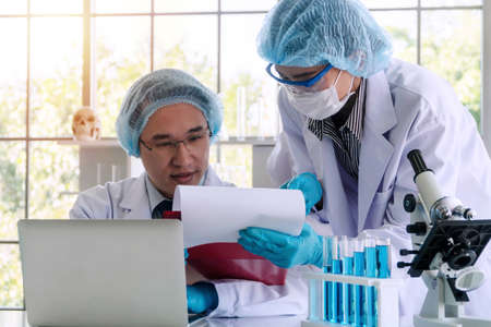 Young asian woman and senior man researcher scientist with laptop, lab coat, gloves and eyeglasses analyzing experiment together in laboratory while looking document and writing note. Science concept.の写真素材
