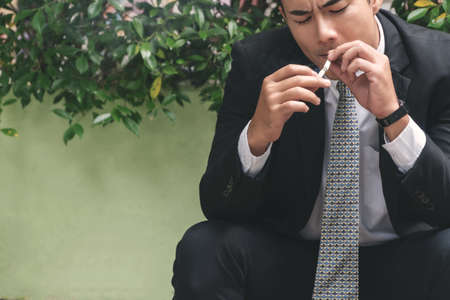 Handsome young businessman wear black suit smoking and thinking about business issues while sitting at outdoor. Young man holding cigarette in his hands while looking at something.の写真素材