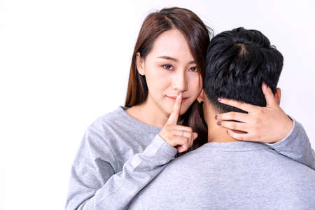 Young women embracing her husband while showing silence sign with forefinger touching secret...