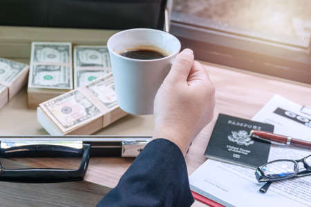 Hands of businessman holding cup of hot coffee with banknote on the bag and glasses , passport,  pen...