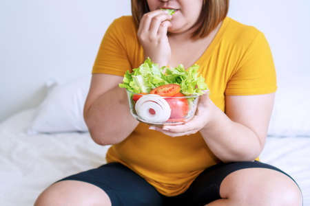 Obesity young women eat green organic vegetables while sitting on the white bed. Fat female holding bowl salad vegetarian while eating lettuce in her hand. Concept food good healthy for weight loss.の写真素材