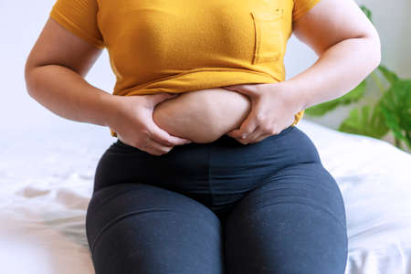 Overweight young woman pinches fat on her belly large size while sitting on white bed at home. Upset female suffering from extra weight. Obesity unhealthily concept.の写真素材