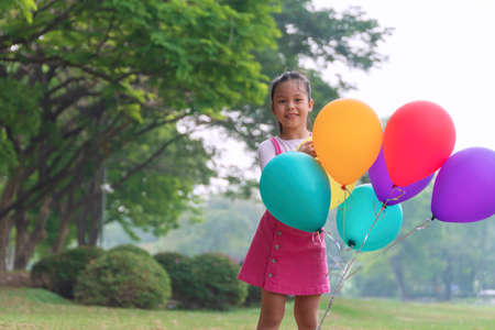 Cheerful children cute girl playful multicolored balloons with big tree on green meadow in the park. Joy kid girl black hair wear pink dress standing with colorful balloons in the garden.の写真素材