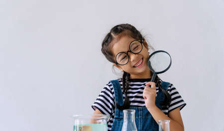 Childhood girl wearing glasses smiling and holding magnifying glass while doing science experiment at table in classroom looking at camera. Innocence kid with test tube front view. Education concept.の写真素材
