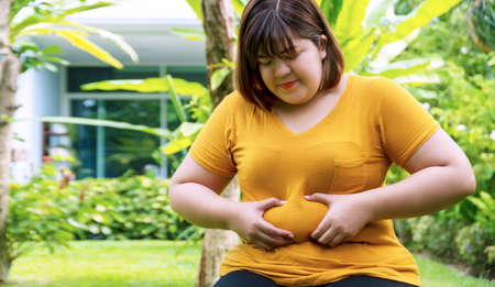 Overweight young woman pinches fat on her belly large size while sitting in the garden at home. Upset female suffering from extra weight. Obesity unhealthily concept.の写真素材