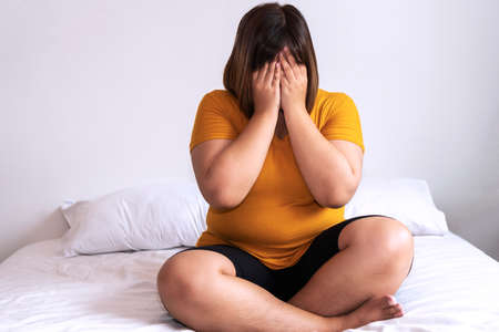 Overweight young woman sitting on white bed while holding hands cover on her face at home. Upset female suffering from extra weight in the bedroom. Obesity unhealthily concept.の写真素材
