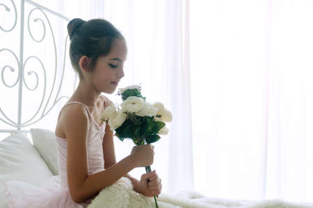 Lovely cute little girl holding bouquet flower in her hand while sitting on the bed with pillow at home. Adorable children wake up in the morning on the white bed fell relax.の写真素材