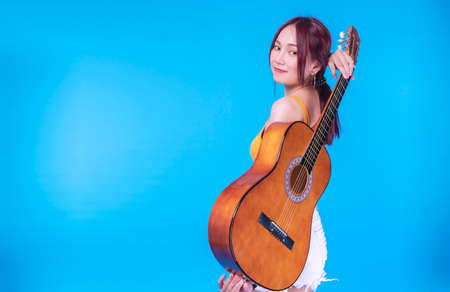 Happiness young woman with an acoustic guitar behind her back on blue background. Portrait cheerful...
