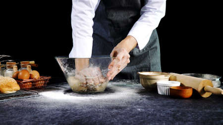 Hand of chef man wear grey apron knead the dough in the glass bowl with egg on table. Ingredients for cooking products or bread, muffins, pizza, pie over black background.の写真素材