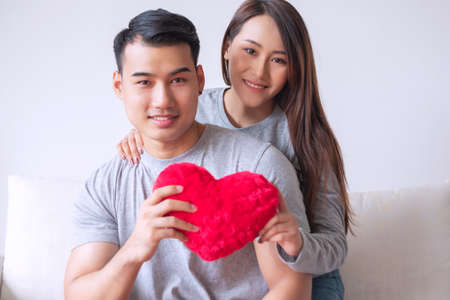 Sweetheart young couple embracing together while holding red heart in their hands at living room. Hands of young happy couple hold red heart cloth symbol love. Valentine day concept.の写真素材