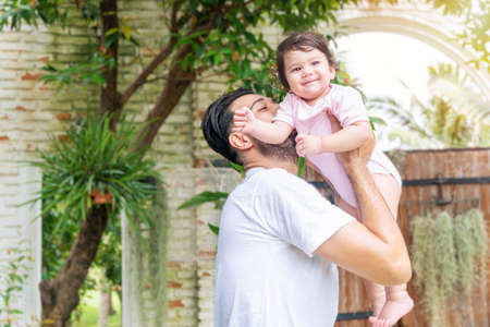 Cheerful father holding and kiss baby daughter in his hand while relax time at home. Mixed race...