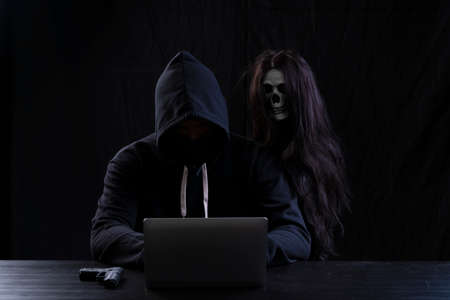 Mysterious man in the hood with skull inside using laptop on table with gun on isolated black...