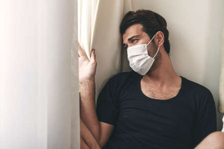 Unhappy young man wear casual cloth and protective face mask worried something while sitting beside windows alone in the room. Quarantine at home concept.の写真素材