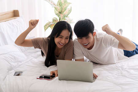 Excited asian couple watching game or winner bid or tv online or exam with laptop while lying on the bed together at home. Lifestyle internet technology and new normal concept.の写真素材