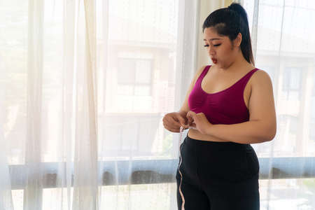 Obesity unhealthy concept. Overweight woman wear sport bar with measure tape on body pinches fat on her belly large size while standing beside windows feeling unhappy.の写真素材