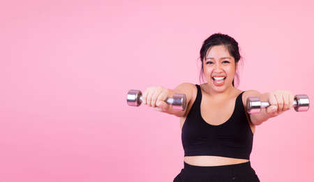 Exercise bodybuilding diet healthy care concept. Chubby obesity plus- size smile woman holding dumbbells workout for weight loss while standing over isolated pink background.の写真素材