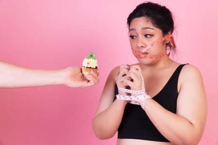 Diet restricts, weight loss, eating disorder concept. Obese overweight fat woman with measure tape cover her mouth while hands of man holding cupcake her thinking eat it over isolated pink background.の写真素材