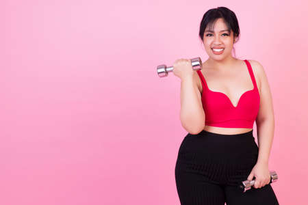Exercise bodybuilding diet healthy care concept. Chubby obesity plus- size smile woman wear sportswear holding dumbbells workout for weight loss while standing over isolated pink background.の写真素材