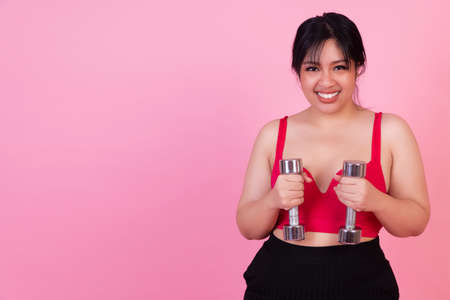 Exercise bodybuilding diet healthy care concept. Chubby obesity plus- size smile woman wear sportswear holding dumbbells workout for weight loss while standing over isolated pink background.の写真素材