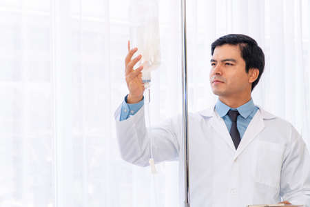 Professional doctor wear white coat uniform checking saline bag for patient at hospital room. Physician handsome young man care drip fluid saline bottle. Health care concept.の写真素材