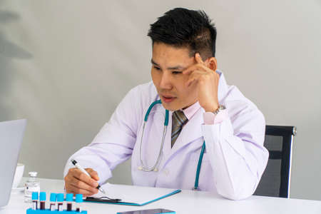 Stress tired doctor wear uniform with stethoscope thinking something while sitting on desk have blood samples test holding pen with folder paperwork in the office clinical. Health care concept.の写真素材