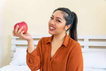Obesity Asian fat woman eating red apple in her hand while sitting on the bed at home. Problem body plus size dieting healthcare with fruit. Weight loss food concept.の写真素材