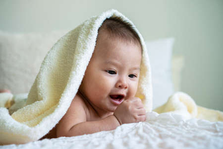 Cheerful cute baby looking at camera under white blanket. Innocence baby crawling on white bed with towel on his head at home.の写真素材