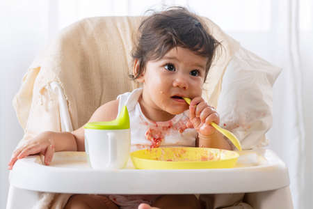 Little cute child celebrating her first birthday with cake at home. Baby adorable girl with apron holding spoon in her hand while sitting on the chair eating cake sloppy her face.の写真素材
