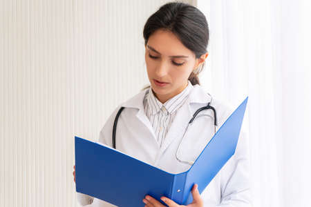 Health care and medical concept. Professional woman doctor wear coat uniform with stethoscope reading information or data disease on paper clipboard while standing beside windows at hospital.の写真素材