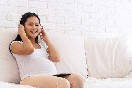 Health care pregnancy concept. Enjoy pregnant young woman wear earphones listening sound music with tablet feeling relaxed while sitting on white sofa at home.の写真素材