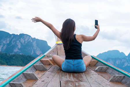 Traveler, vacation, summertime in holiday concept. Enjoy Asian woman using smartphone selfie photo feeling freedom and relax while sitting on the boat at tropical sea.の写真素材